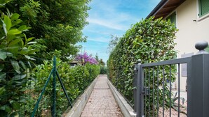 En smal brostenssti omkranset af frodig beplantning og et metalhegn, der fører hen til en bygning med en lys facade og grønne skodder.
