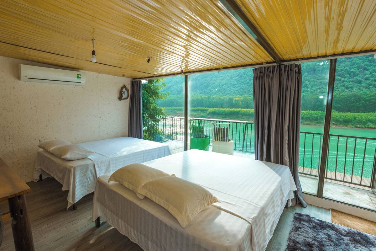 Una habitación con dos camas, ventilador de techo, aire acondicionado y un amplio ventanal con vistas a un paisaje verde.