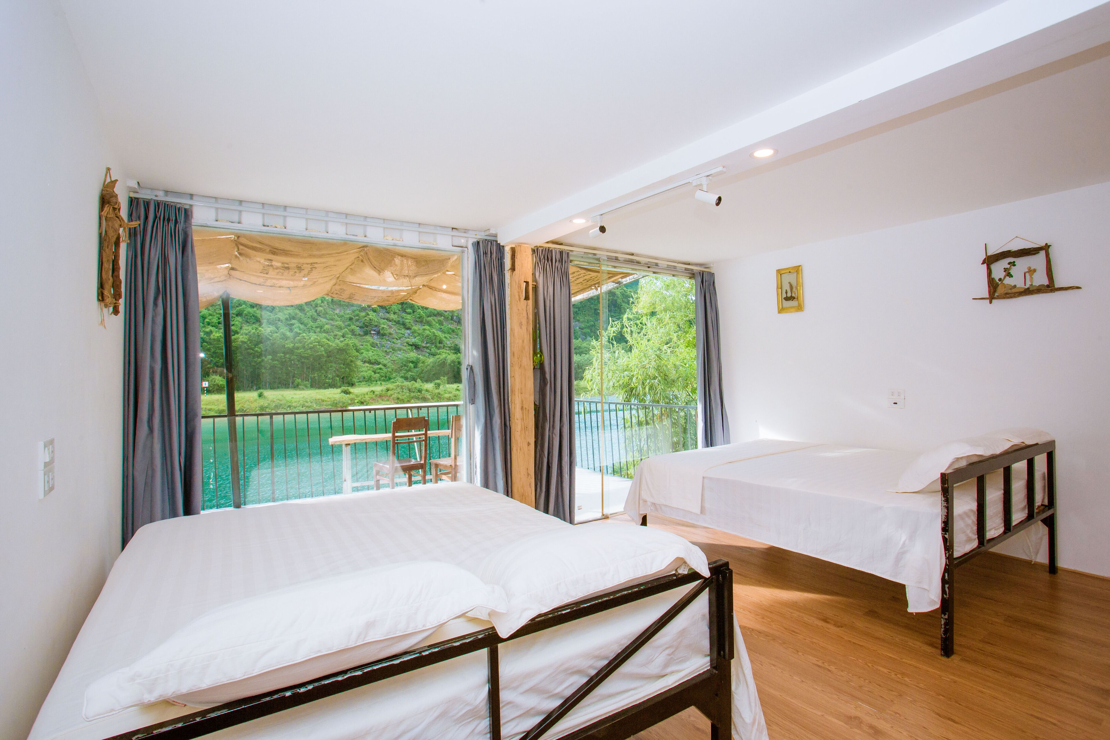 A room with two beds, large windows, and a balcony with a view of greenery.