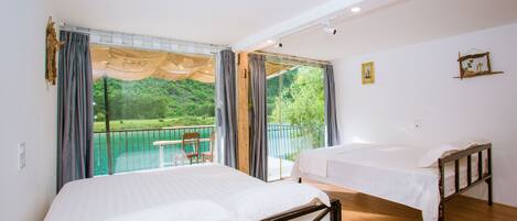 A room with two beds, large windows, and a balcony with a view of greenery.