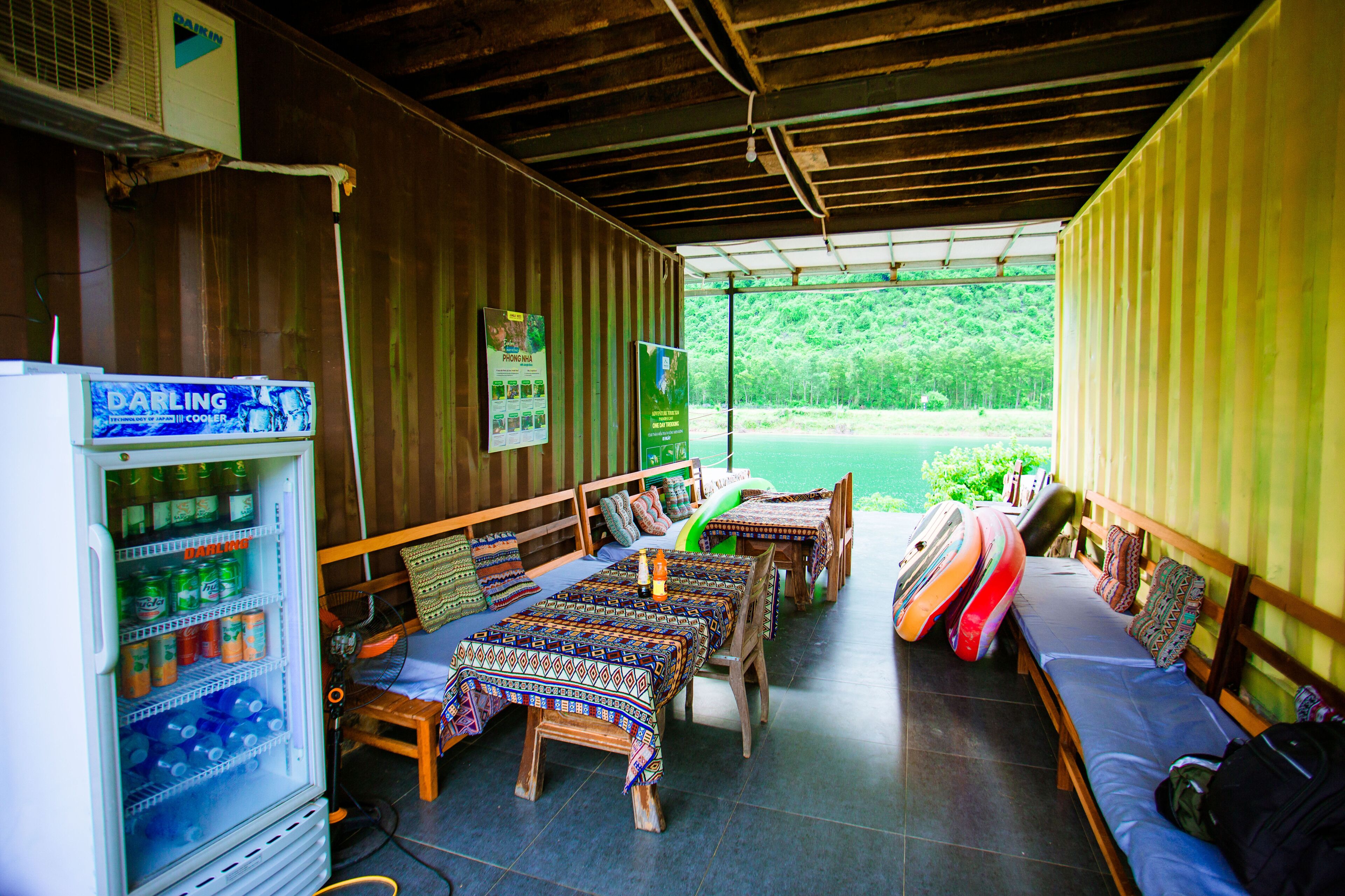 A covered outdoor seating area with tables and benches, a beverage vending machine, and a view of a forested area.
