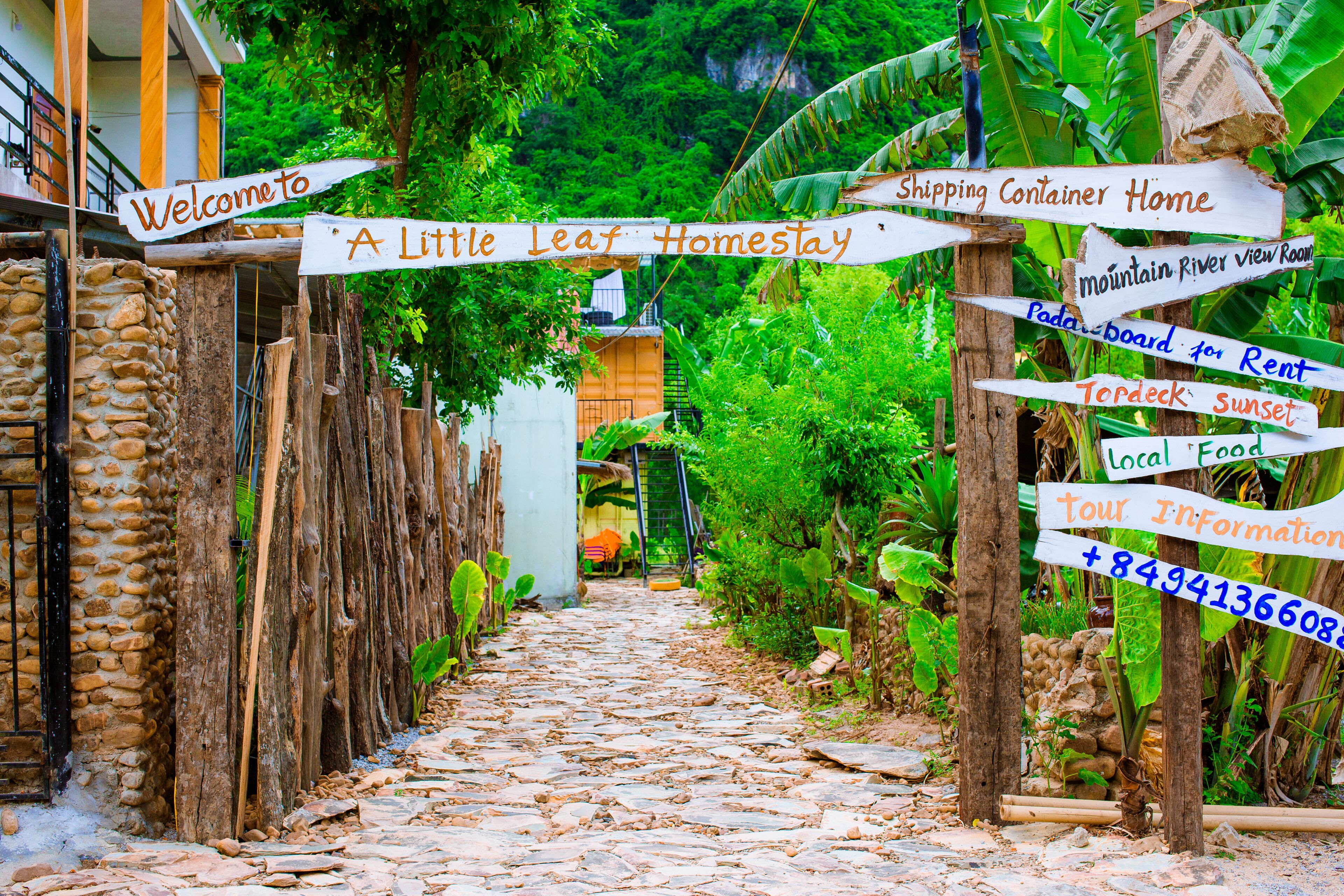 A narrow stone pathway leading to a rustic homestay with signs indicating amenities like a mountain river view room, a paddleboard for rent, and a sunset deck.