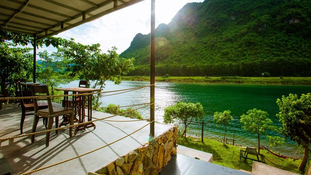A terrace with a table and chairs overlooking a lake, surrounded by greenery and mountains. - A Little Leaf Homestay  (Phong Nha)