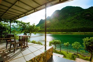 A terrace with a table and chairs overlooking a lake, surrounded by greenery and mountains. - A Little Leaf Homestay  (Phong Nha)
