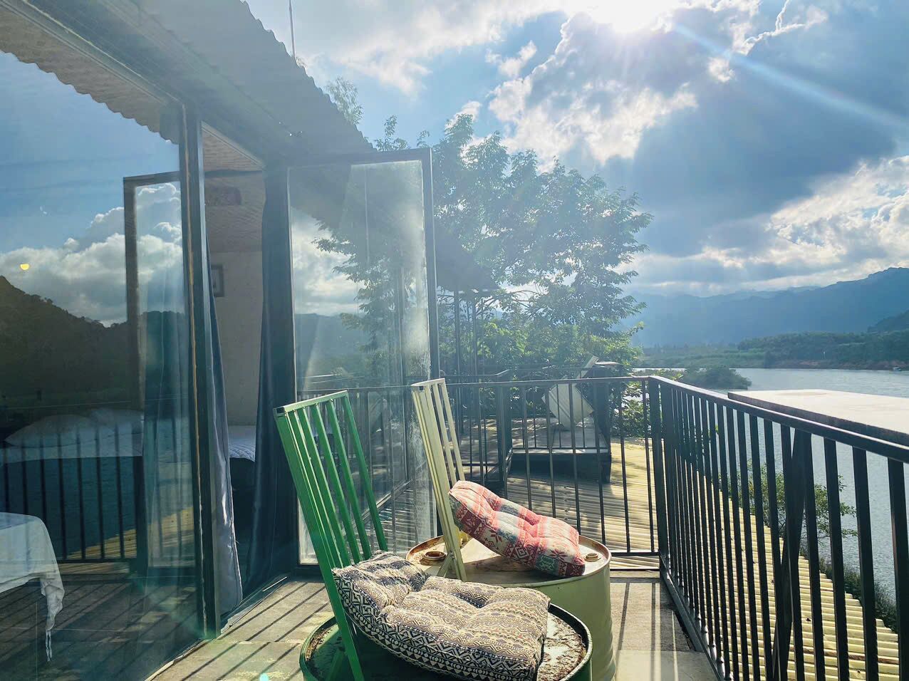 A balcony with a view of a river and mountains, a green chair, a patterned cushion, and a table with a glass enclosure.