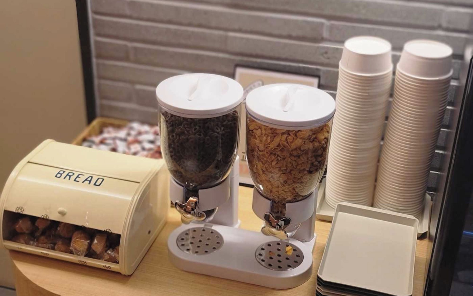 A food preparation area with a bread dispenser, two coffee grinders, and stacks of paper plates.