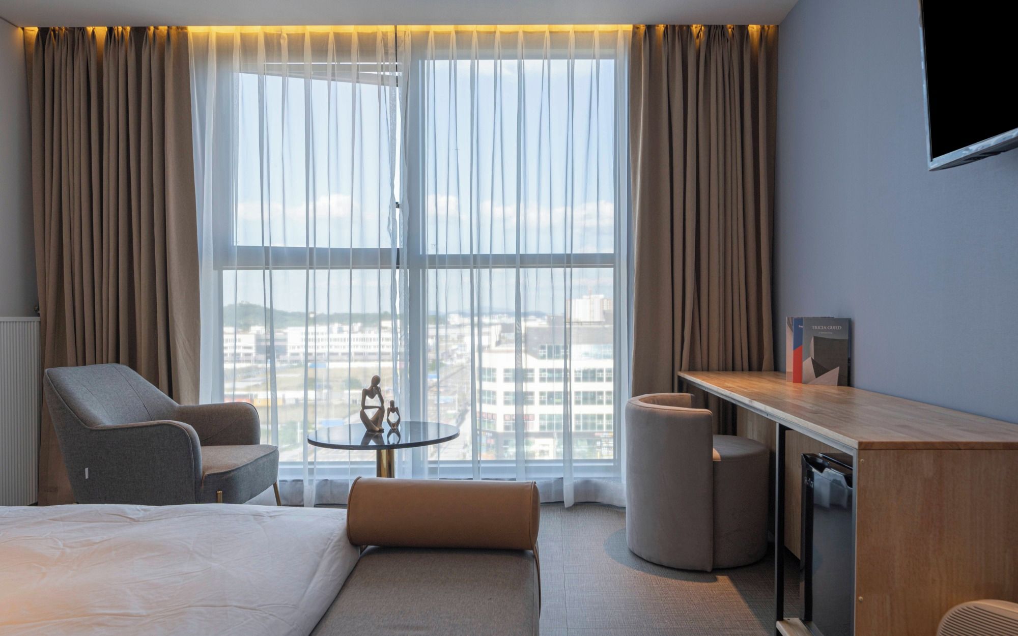A hotel room with a bed, a desk, a chair, a small table, and a view of buildings through large windows.
