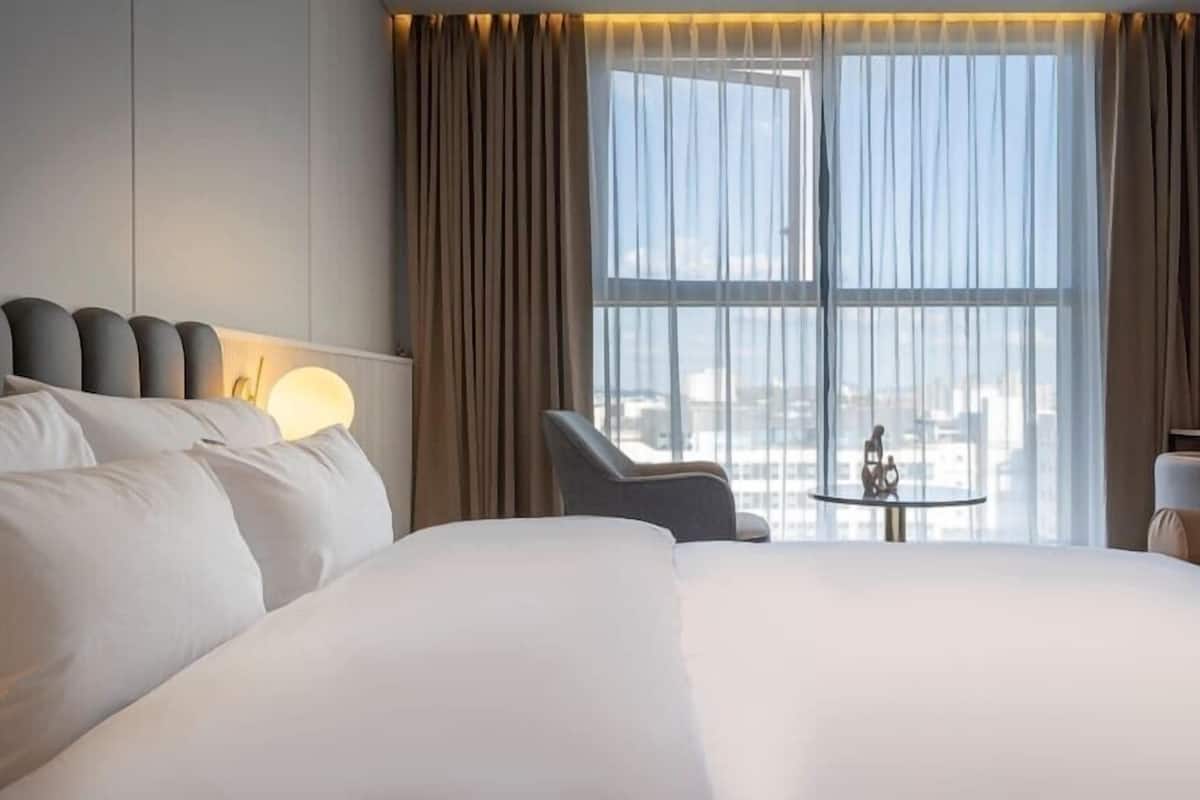 A hotel room with a large bed, a chair, a small table, and a view of the city through large windows.