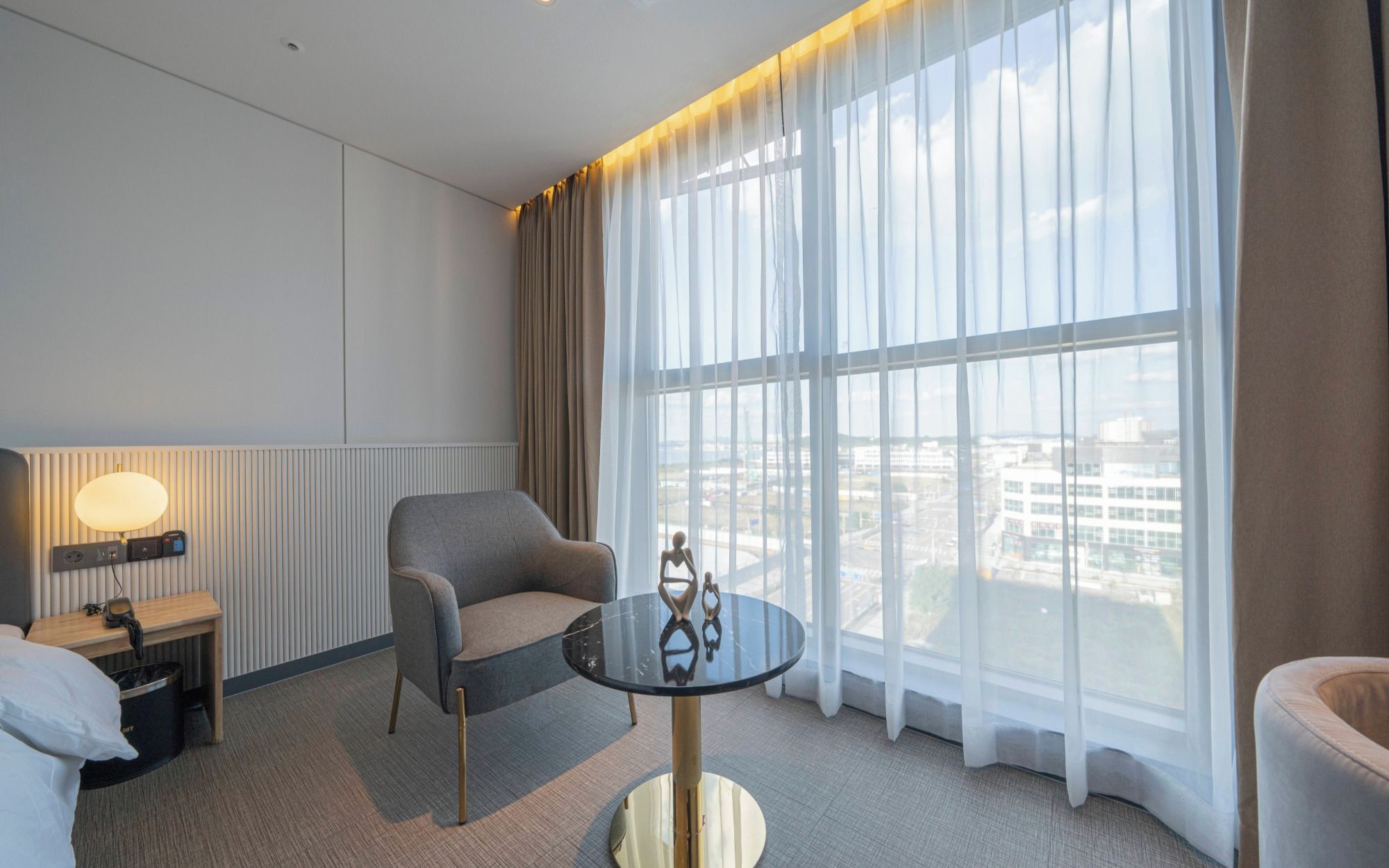 Une chambre d’hôtel dotée d’une grande fenêtre, d’une table ronde, d’une chaise, d’une table de chevet avec une lampe et d’une prise de courant fixée au mur.