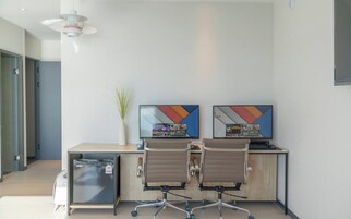Un espacio de oficina moderno con dos escritorios, sillas, una planta pequeña y un televisor de pared que muestra títulos de juegos.