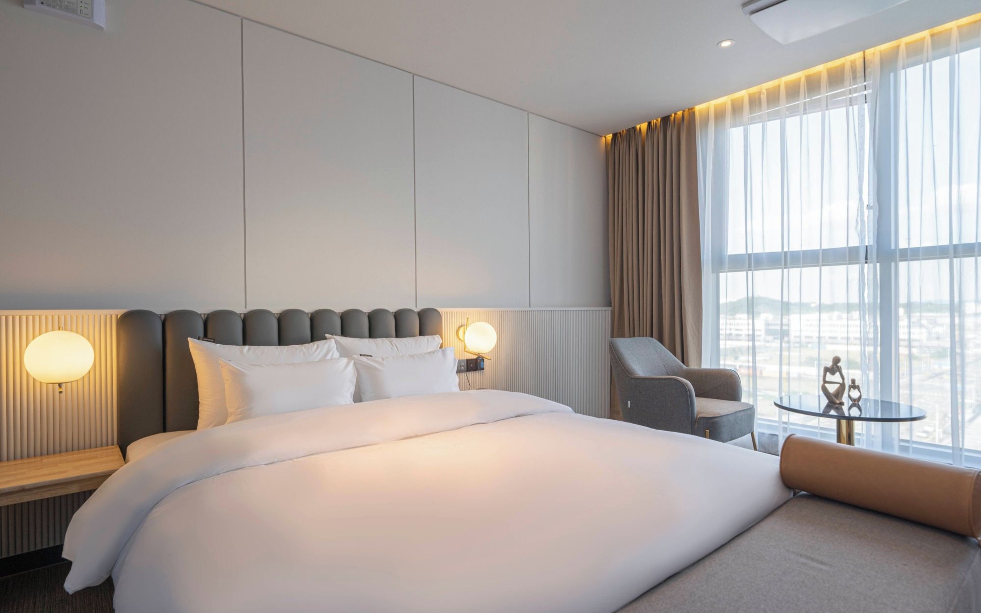 A hotel room with a large bed, a seating area, a small table, and a view of the city through large windows.