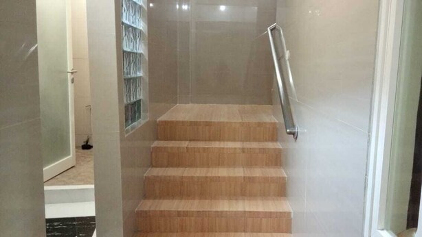 A staircase with wooden steps and a metal handrail, leading to a room with a tiled floor and a door. - BLOOMINGTON (Surabaya)