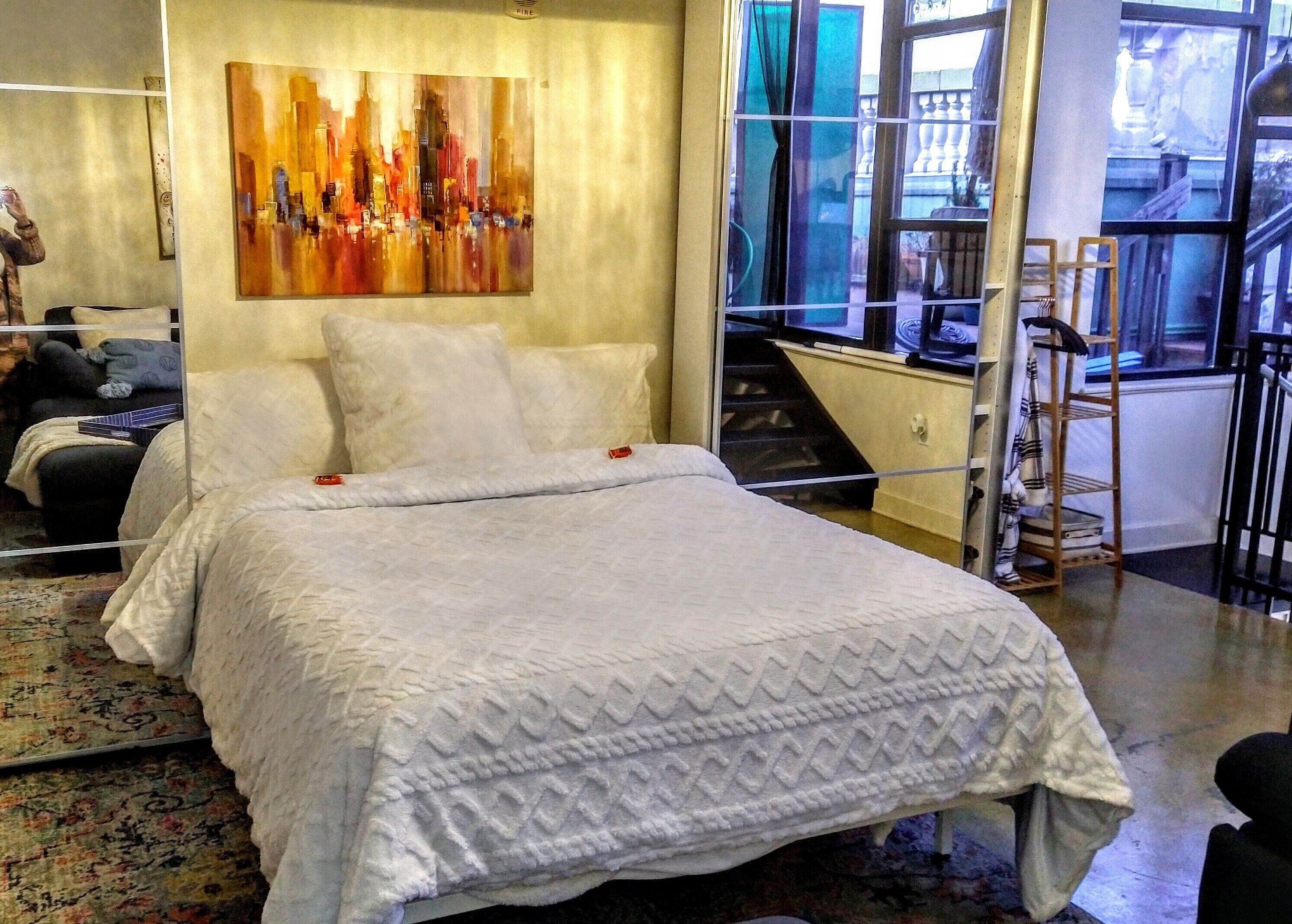 Ein Schlafzimmer mit einem großen Bett, einem Stadtbild an der Wand und Blick ins Freie durch ein Fenster.