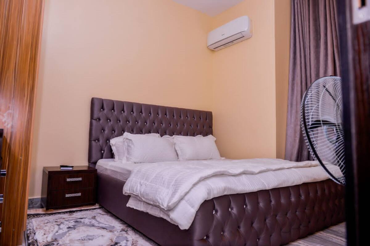 A neatly made bed with a tufted headboard, a bedside table, and a wall-mounted air conditioner.