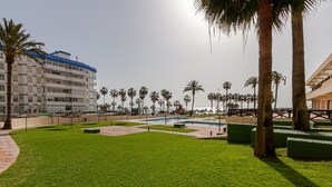 Zona de piscina con palmeras, un edificio con balcones y un cielo despejado.