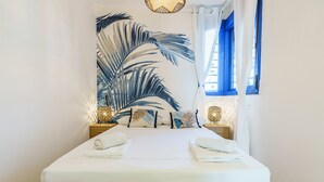 Un lit bien fait, avec du linge de lit et des serviettes blanches, une grande œuvre d’art représentant une feuille tropicale accrochée au mur, et une fenêtre avec des rideaux bleus donnant sur des immeubles.