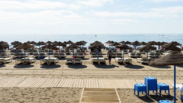 Une plage bordée de rangées de chaises longues et d’ombrelles en chaume, une promenade en bois et une vue dégagée sur la mer.