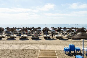Ein Strand mit Liegestuhlreihen und reetgedeckten Sonnenschirmen, einer Holzpromenade und freiem Blick auf das Meer.