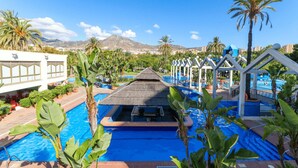 A pool area with a thatched-roof structure, palm trees, and a clear blue sky. - Seaview beachfront studio with aqua park & balcony REF007 (Benalmádena)