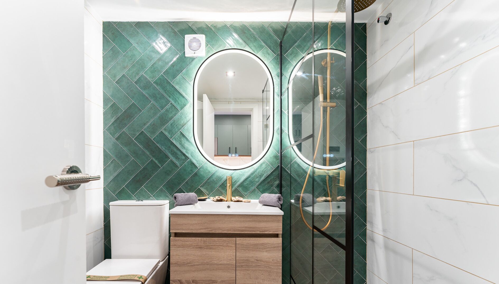 Een badkamer met een groen betegelde muur, een wit wastafel, een houten wastafelmeubel en een douchecabine met glazen wanden.