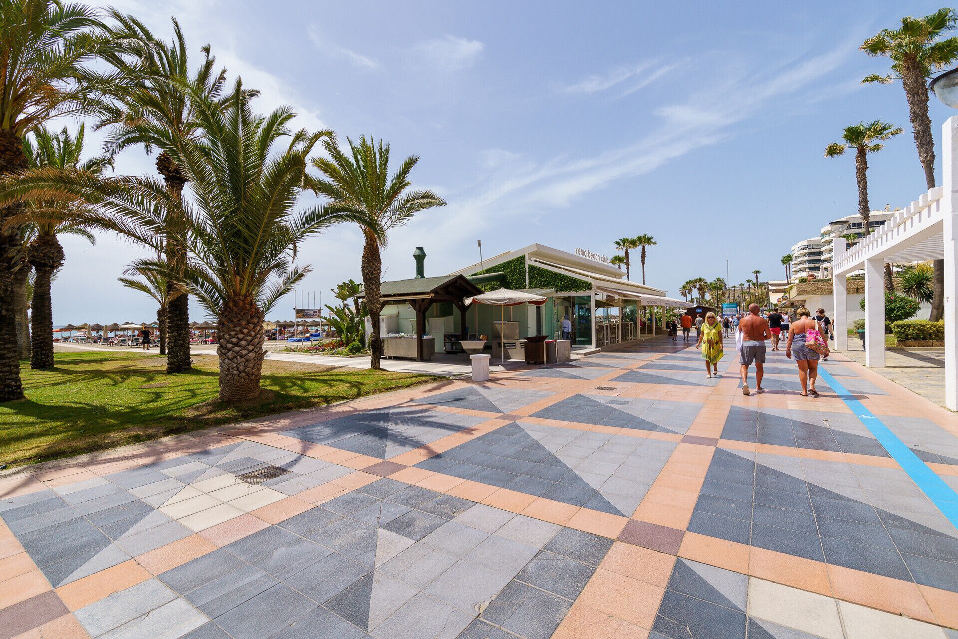 Eine Promenade mit Palmen, einem Beachclub und einem Kieselweg.