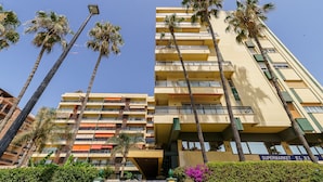 Un hotel de varios pisos con balcones, palmeras y un letrero visible de un supermercado.