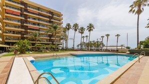 Una piscina con solárium empedrado, rodeada de palmeras y un edificio de varios pisos con balcones.