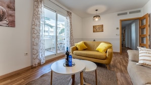 Ein Wohnzimmer mit einer Couch, zwei kleinen Tischen, einem großen Fenster mit Vorhängen, einem Gemälde und einer Tür zu einem anderen Raum.