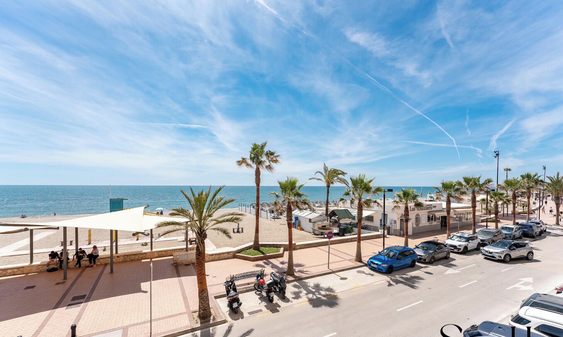 A coastal street with parked cars, palm trees, and a beach in the background.