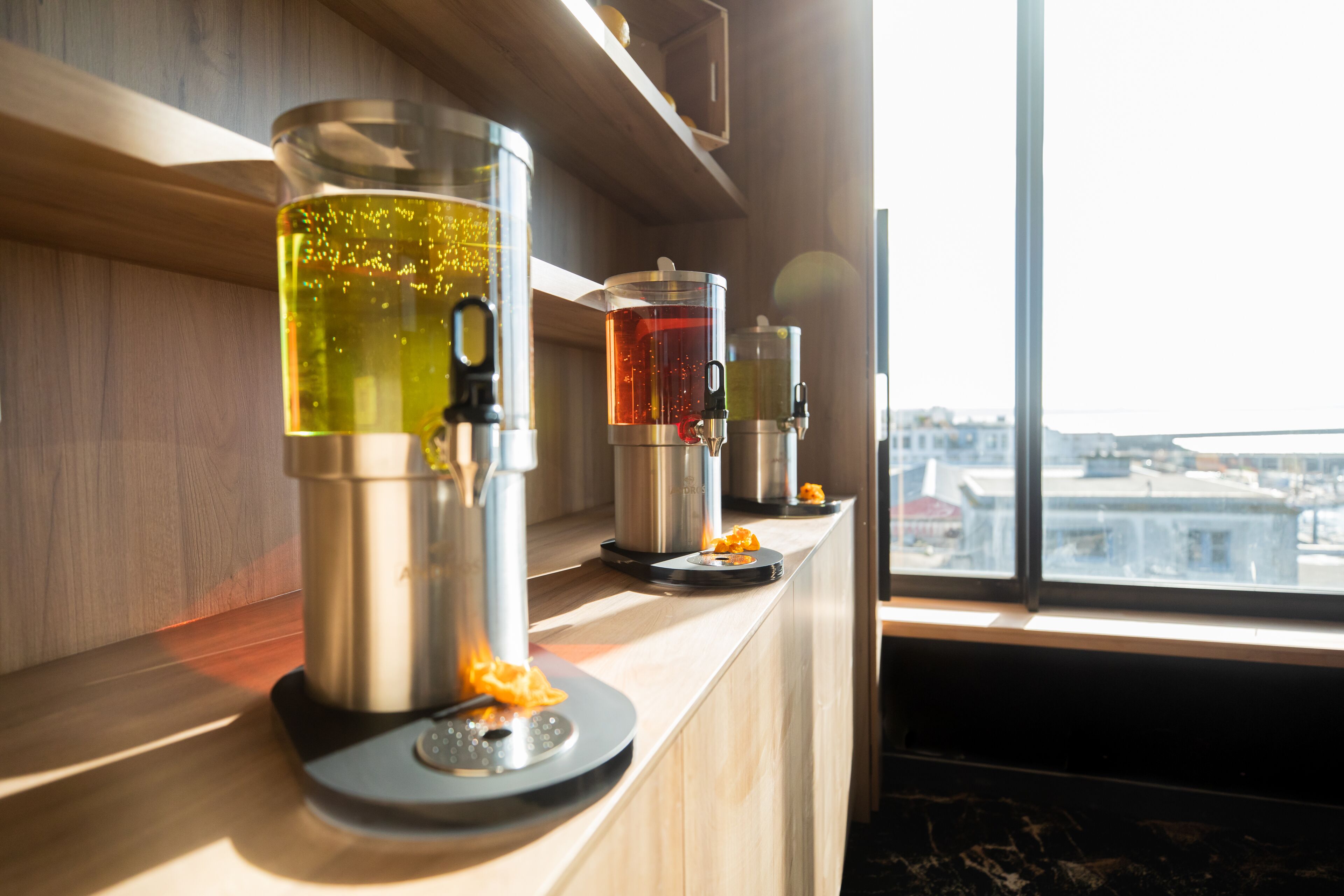 A coffee machine and a tea dispenser on a wooden counter by a window.