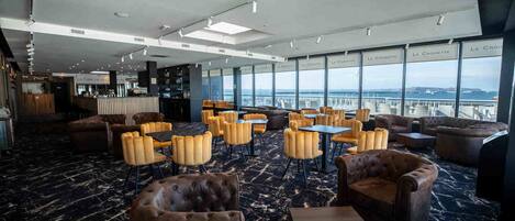 Un amplio salón con sofás de cuero y sillones amarillos, ventanas grandes con vistas a la ciudad, y un área de bar al fondo.
