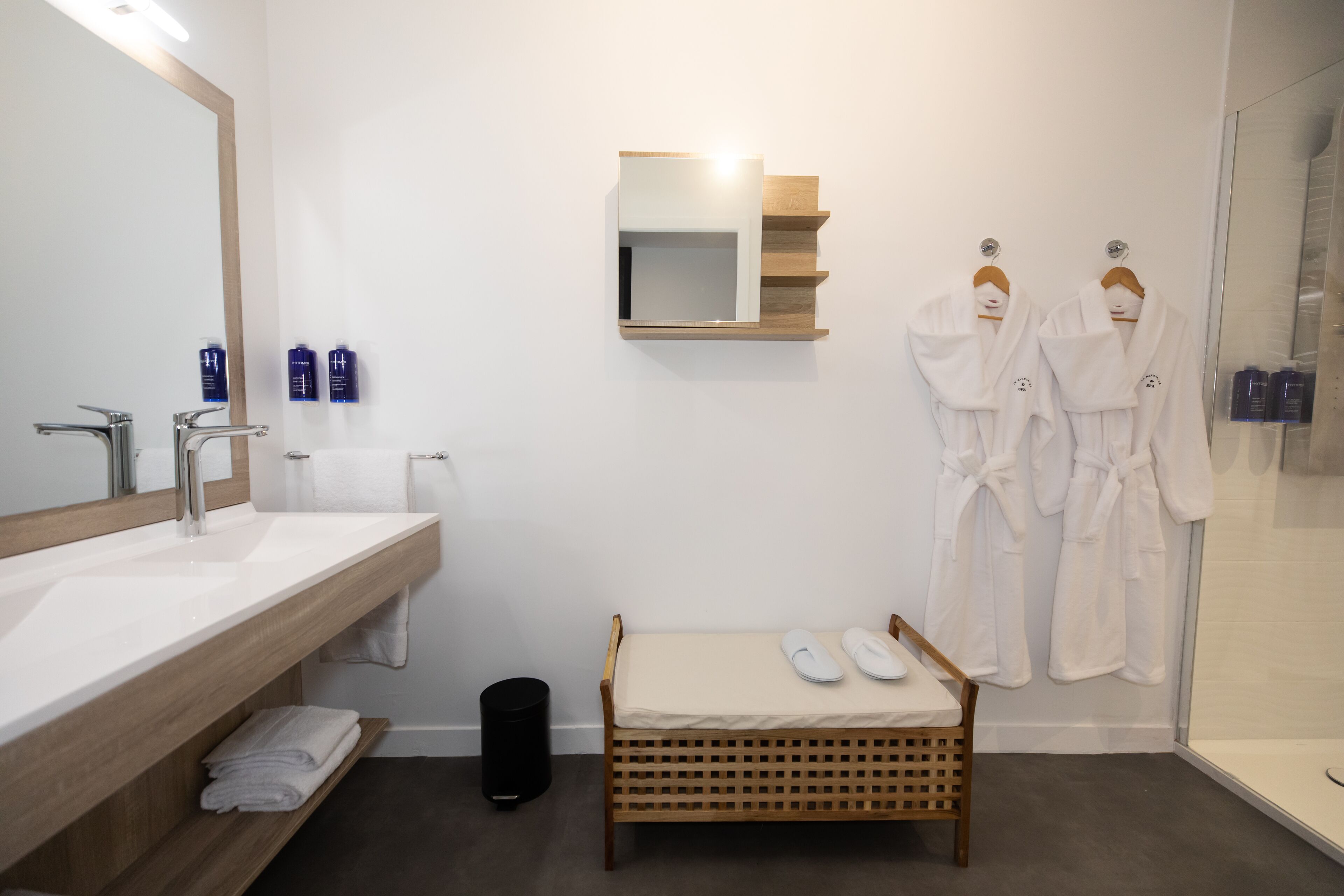 A modern bathroom with a wooden vanity, a large mirror, two white robes hanging, and a small wooden bench with slippers.