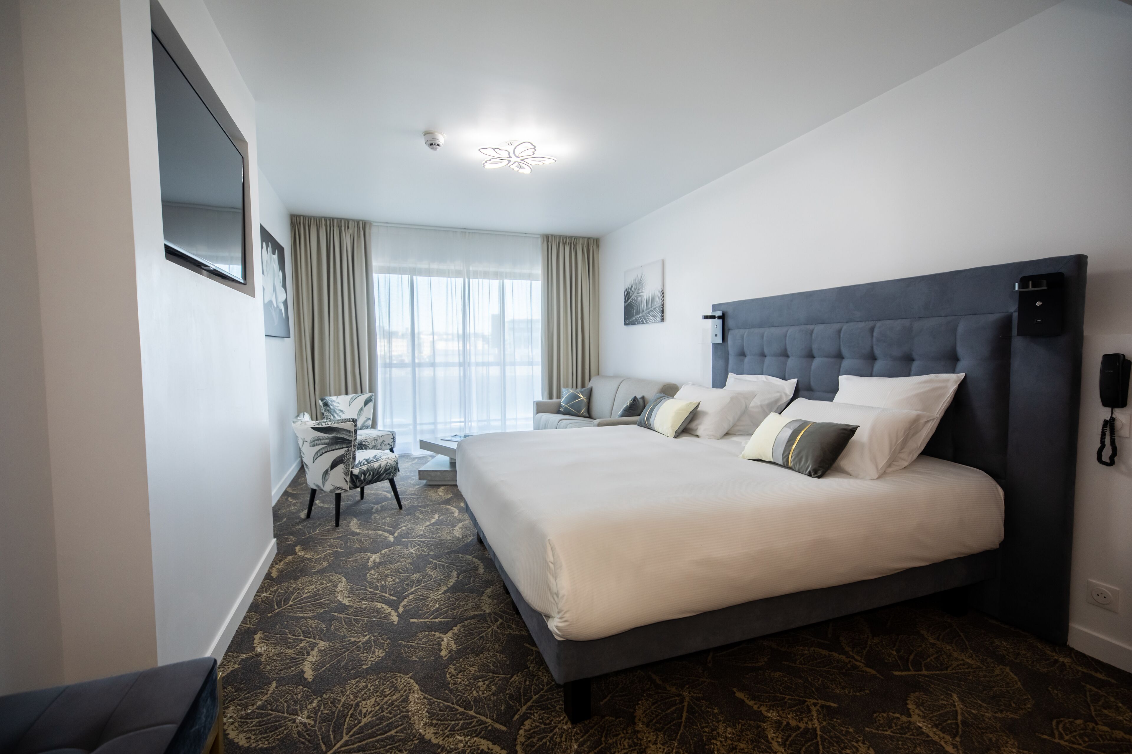 Une chambre d’hôtel moderne dotée d’un grand lit, d’un coin salon avec un canapé et des fauteuils, d’un téléviseur à écran plat fixé au mur et d’une grande fenêtre avec des rideaux.
