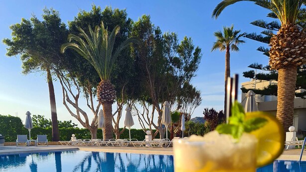 A clear pool with lounge chairs and umbrellas, a tropical setting with palm trees, and a cocktail glass with a mint garnish. - Abella Hotel (Chania)