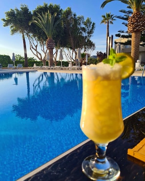 A clear pool with lounge chairs and umbrellas, a tropical setting with palm trees, and a cocktail glass with a mint garnish. - Abella Hotel (Chania)