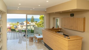 A modern hotel reception area with a wooden desk, a small seating area, and a view of the sea through large glass doors. - Abella Hotel (Chania)