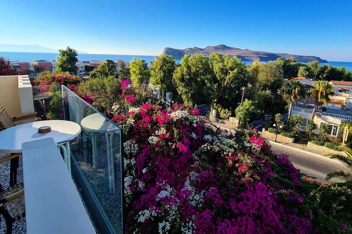 A balcony with a table and chairs, overlooking a coastal view with bougainvillea flowers and a clear blue sky.
