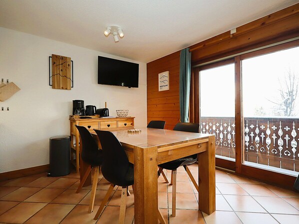 A dining area with a wooden table and chairs, a television mounted on the wall, and a window with a view of the outdoors.