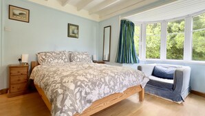 Un dormitorio con cama, mesitas de noche, una silla, una ventana con cortinas y dos cuadros en la pared.