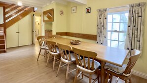 Un comedor con mesa y sillas de madera, una escalera y un ventanal con cortinas.