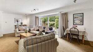 A living room with a sofa, a coffee table, and a dining table with a chair. There is a window with curtains, a radiator, and a framed picture on the wall. - Loughrigg -  a bungalow in the centre of Rock with parking (Rock)