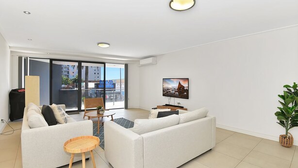 En moderne stue med sofa, et lille bord, fjernsyn og en balkon med udsigt til bygninger.