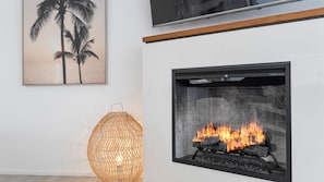 A modern living room with a fireplace, a framed picture of palm trees, and a woven floor lamp.