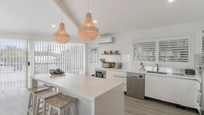 A modern kitchen with white cabinetry, a central island, pendant lighting, and a view through the glass door. - Sunset Lake House - Pool, Lake Views & Beach (The Entrance North)