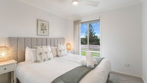A bedroom with a large bed, a headboard, a bedside table with a lamp, a framed picture on the wall, and a window with a view of trees and a road. - Sunset Lake House - Pool, Lake Views & Beach (The Entrance North)