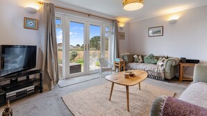 Ein Wohnzimmer mit Fernseher, Couchtisch, Sofa und Blick durch große Glasschübe nach draußen.
