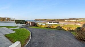 Zona residencial com calçada pavimentada que leva a uma casa de design moderno, um pequeno jardim e vista para o mar e colinas ao fundo.