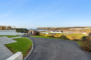 Ein Wohngebiet mit befestigter Auffahrt zu einem Haus im modernen Stil, einem kleinen Garten und Blick auf das Meer und die Hügel im Hintergrund.