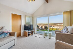 Ein Wohnzimmer mit Fernseher, zwei Sofas, einem Glastisch und einer Glasschiebetür, die auf eine Terrasse führt.