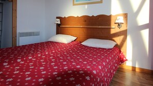 Een bed met een rode bloemenprint deken, twee witte kussens, een houten hoofdbord en een lijst met een foto van een besneeerd landschap aan de muur.